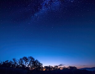 A serene view of a star-filled sky over a silhouette of trees and hills. Dark blue gradient sky filled with stars and a light source far away