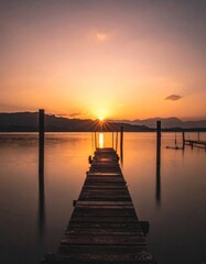 Obraz premium A wooden pier leads into calm, reflective water beneath a vibrant orange sunset over distant mountains, creating a tranquil atmosphere