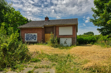 Wide shot of deteriorating brick house with boarded windows and graffiti under cloudy sky. Overgrown grass and vegetation surround the abandoned structure.