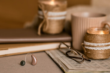 Behind-the-ear hearing aid lying near warm candles, a notebook, and reading glasses. Hearing...