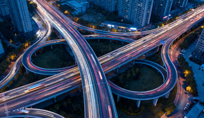 城市立交桥航拍，多层环形匝道如金属丝带缠绕，车流形成彩色光轨，周边高楼环绕，黄昏蓝调时刻 © stay