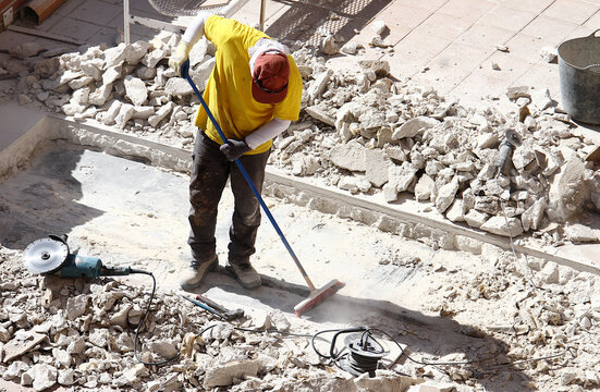 Obrero barriendo y limpiando con escoba el polvo entre piedras, escombros, radial, cubo y cables en obra reforma de suelo del patio