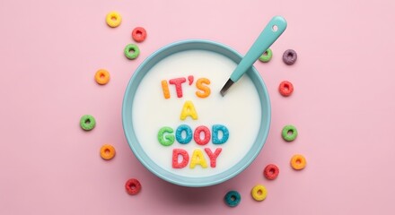 Colorful cereal letters spelling its a good day floating in a bowl of milk with a spoon and scattered around on a pink background
