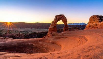 Delicate Arch stands silhouetted in the sunset with mountains on horizon, glowing orange in the light