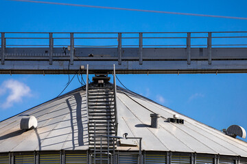 Fototapeta premium A high-angle shot shows several large, silver grain silos under a partly cloudy blue sky. Metal walkways and structures connect the silos. The scene suggests an industrial agricultural facility.
