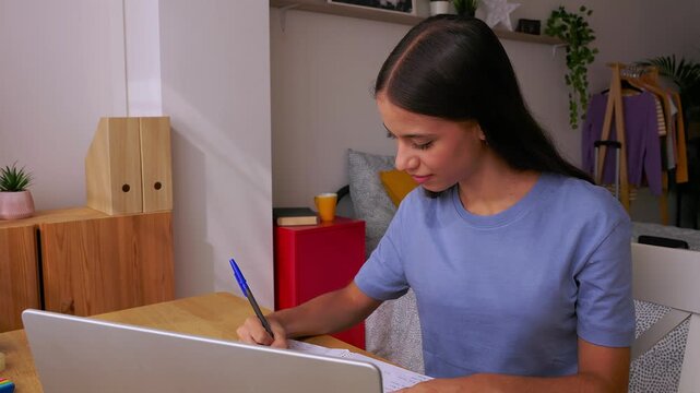 15 years old teenage student girl studying online with laptop at home. Focused teen doing homework at desk with notebooks and headphones nearby. Education and e-learning lifestyle concept.