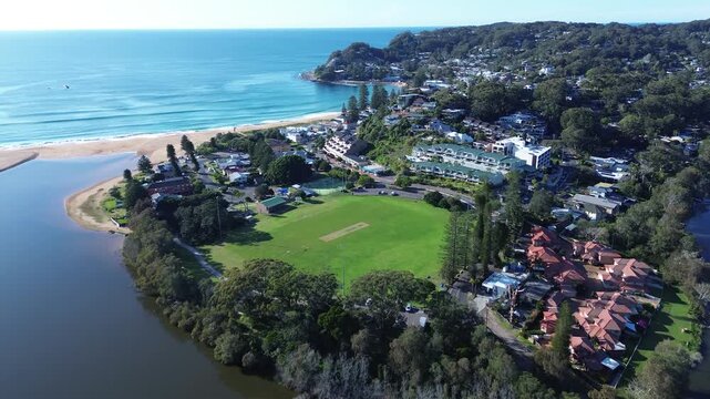 Drone aerial of Avoca Beach public sports oval Heazlett Park surrounded by residential housing town shops and tree-lined streets with ocean coastal waves breaking in background Central Coast Australia