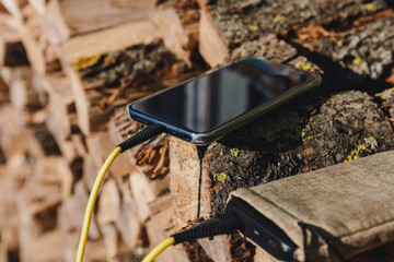 Smartphone charging with a powerbank on a wooden log pile in a lush outdoor setting. The bright yellow cable highlights the tech amidst the natural backdrop, ideal for outdoor tech themes.