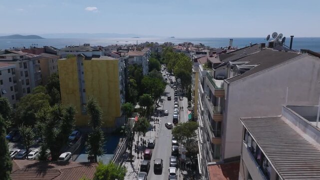 Aerial view of a coastal city with red-roofed buildings, a seaside park, and a wide view of the sea and skyline under a cloudy sky.