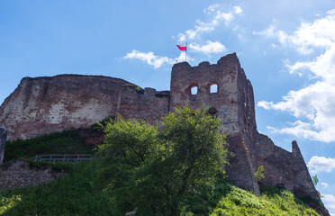 Fototapeta premium The stone ruins of a medieval castle tower rise against a blue summer sky, with the Polish national flag waving proudly on top. A historical landmark symbolizing cultural heritage and centuries of his