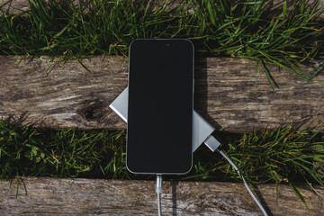 The smartphone is charged by a power bank on a rustic wooden surface surrounded by green grass.