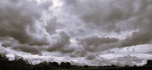 A sweeping sky of dark, textured clouds rolls over a silhouetted landscape. The grayscale palette...