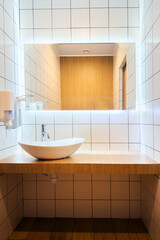 Contemporary minimalistic design indoors bathroom with white oval sink on wooden counter top and chrome faucet. Tile wall with mirror and warm light and soap dispenser. Vertical view