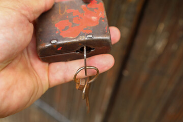 ​A hand holds an old, rusty lock with damaged red paint, from which a bunch of keys hangs. Light reflects off the metallic surface, highlighting the age and wear of the object.