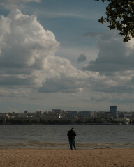 A view of a cityscape across a river or body of water on an overcast day. In the foreground, on a sandy shore, stands a lone person. Thick clouds in the sky create a pensive atmosphere.