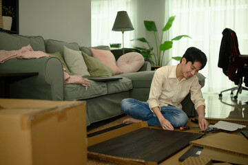 Fototapeta premium Indoor lifestyle scene of a man sitting cross-legged, unpacking furniture parts and arranging tools for assembly