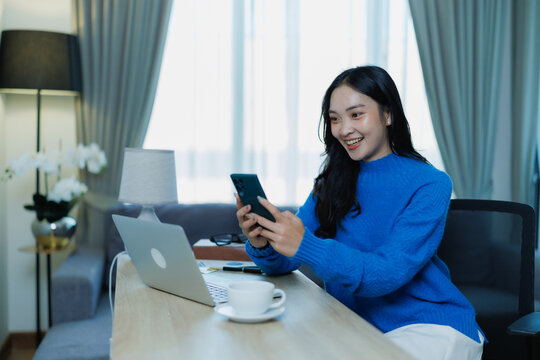 Young Asian woman smiling while working from home on a laptop and smartphone, connecting with colleagues via video call and messages in a cozy living room office space