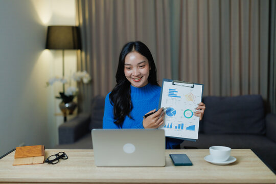 Smiling young Asian woman presenting financial charts and graphs on a clipboard during a remote online meeting or video conference call from her home office, communicating effectively - Powered by Adobe