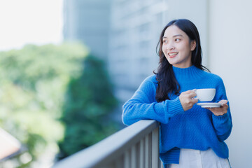 Asian woman standing on a balcony in a cozy sweater, smiling and holding a cup of coffee while enjoying a peaceful morning, fresh air and quiet urban relaxation