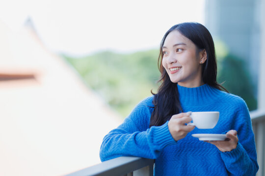 Young Asian woman stands on a balcony in a blue sweater, holding a cup and saucer, smiling contentedly as she enjoys fresh morning air and a peaceful, relaxing moment - Powered by Adobe
