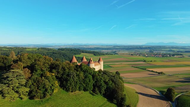 Aerial view of Champvent Castle nestled amidst lush greenery contrasting with the patchwork fields under a clear blue sky, Champvent, Vaud, Switzerland.