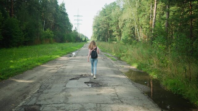 Young girl with loose hair walking thoughtfully on tarred forest road with potholes and puddles, surrounded by lush greenery and tall trees, soft daylight casting gentle shadows