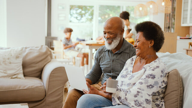 Senior Couple At Home Looking At Laptop Together With Multi-Generation Family In Background