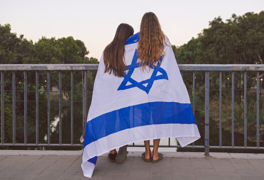 Two Israeli teen girls wrapped in the flag of Israel, looking over a bridge