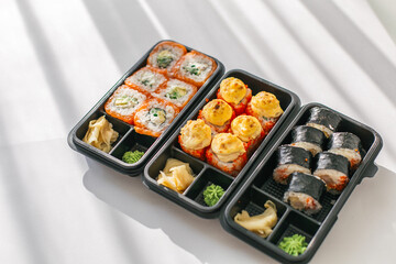 Assorted sushi rolls in black plastic takeaway containers on white background with natural sunlight. Japanese cuisine with fresh rolls, baked sushi, wasabi, and pickled ginger for delivery or takeout