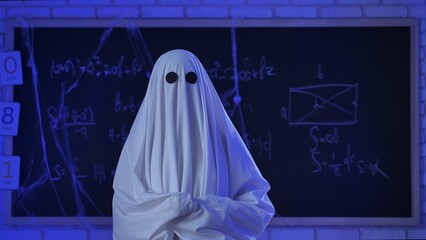 A person in a white ghost sheet sits near the blackboard and teaches a class in a blue lit classroom