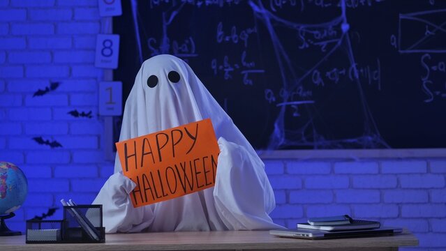 A person dressed in a white ghost sheet holds a Happy Halloween sign in a classroom with blue lighting - Powered by Adobe