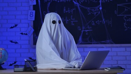 A person in a white ghost sheet sits at a table in a blue-lit room and types on a laptop