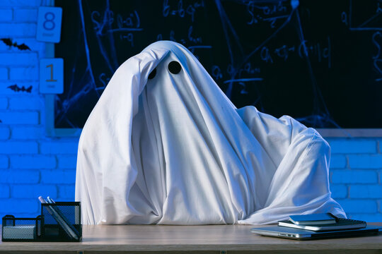 A man in a white ghost sheet sits at a table in a blue-lit room and clutches his head in despair