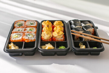 Variety of sushi in black plastic containers with chopsticks on a white table. Traditional Japanese food with baked rolls, fresh rolls, wasabi, and pickled ginger