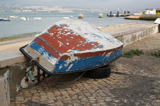 Overturned boat on cobblestones near the waterfront in Trafaria, Portugal. Weathered hull, rope, tire, and boats on the water create a composition of texture, contrast, and maritime character.