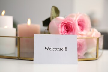 Card with word Welcome, roses and burning candles on white table, closeup