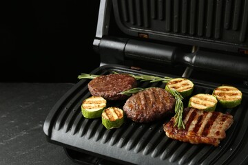 Electric grill with beef steaks, zucchini and rosemary on black table, closeup. Space for text