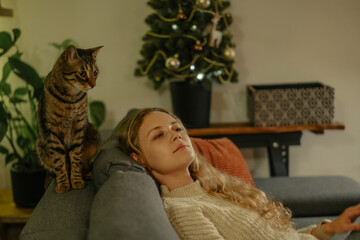 Young cute blonde woman relaxing with her tabby cat at home on the couch, Christmas holidays with pet