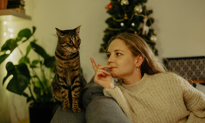 Young cute blonde woman relaxing with her tabby cat at home on the couch, Christmas holidays with pet