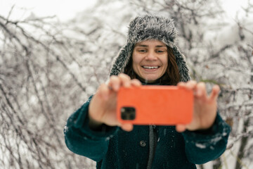Young woman enjoying snowfall and using smartphone while walking in winter woodland