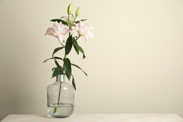 Beautiful lily flowers in glass vase on table near beige wall, space for text