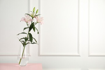 Beautiful lily flowers in glass vase on pink wooden table near white wall indoors, space for text