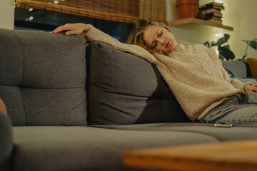 Relaxed young woman in warm sweater lying on sofa at home in evening