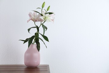 Beautiful lily flowers in vase on wooden table near white wall indoors, space for text