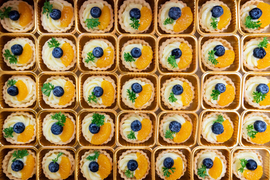 Top view of assorted mini fruit tarts in golden trays, decorated with blueberries, orange slices. Colorful and elegant dessert presentation, perfect for catering, bakery menus, or food blogs.