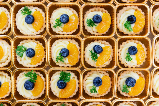 Top view of assorted mini fruit tarts in golden trays, decorated with blueberries, orange slices. Colorful and elegant dessert presentation, perfect for catering, bakery menus, or food blogs.