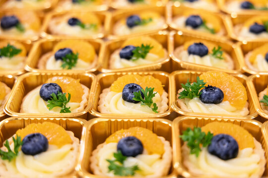Close-up of assorted mini fruit tarts in golden trays, decorated with blueberries, orange slices. Colorful and elegant dessert presentation, perfect for catering, bakery menus, or food blogs.