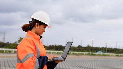 A female construction worker wearing a hard hat and safety jacket is focused on her laptop at a construction site. The sky is overcast, indicating possible rain