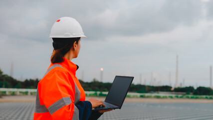 A construction engineer wearing a safety helmet and bright orange jacket reviews data on a laptop at a solar installation site. The sky is overcast, indicating a chance of rain