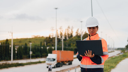 A construction worker observes traffic and analyzes data on her laptop along a transport route during sunset. The scene captures a blend of nature and industry